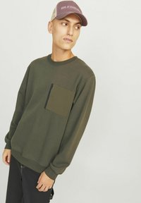 Olivgrüner Sweatshirt mit einer vorderen aufgesetzten Tasche in dunklerem Grün, Rundhalsausschnitt und langen Ärmeln, kombiniert mit dunklen Hosen.