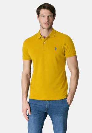 Uomo in piedi con le mani parzialmente nelle tasche, indossa una polo giallo senape e jeans blu su uno sfondo semplice.