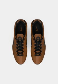 Καφέ αθλητικά παπούτσια με φάρδυ ανώτερο τμήμα από δίχτυ και συνθετικό υλικό, μαύρες λεπτομέρειες, στρογγυλά κορδόνια και χαμηλή σχεδίαση με εμφανές λογότυπο της Nike.