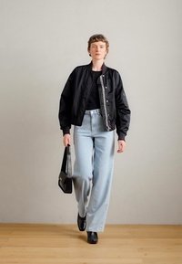 Μαύρο μπόμπερ μπουφάν με φερμουάρ, συνδυασμένο με ανοιχτό γαλάζιο παντελόνι φαρδιάς γραμμής. Το μοντέλο κρατά μια μαύρη τσάντα και φορά μαύρες μπότες μέχρι τον αστράγαλο.