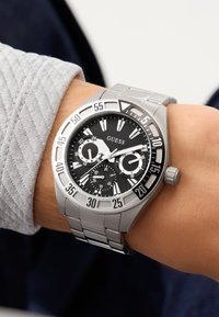 Pánské stříbrné kovové hodinky značky Guess se černým ciferníkem, třemi subciferníky a velkými minutovými značkami nošené na levém zápěstí s šedým rukávem.