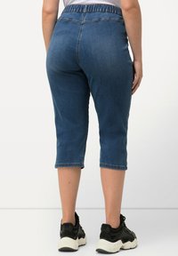 Pantalones capri de mezclilla con cinturilla elástica, en un tono azul oscuro, con corte recto y longitud recortada a la altura de la pantorrilla. Combinados con zapatillas negras.