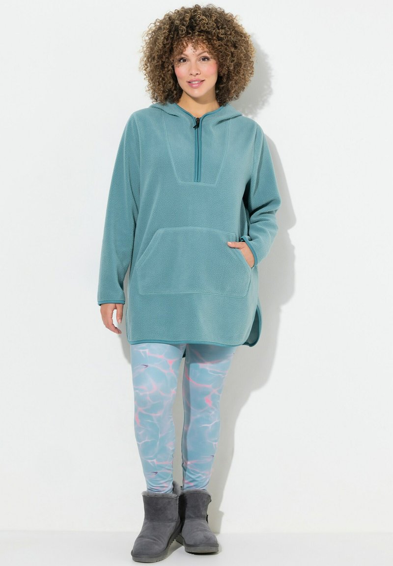 Teallin fleece pullover, jossa on puolivetoketju, kenguru-tasku ja sivuaukot, yhdistettynä vaaleansinisiin legginsseihin, joissa on vesiaallokku-kuvio ja harmaat saappaat.
