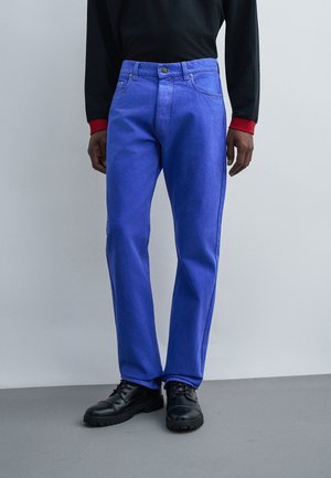 Personne portant un jean bleu vif à jambes droites, des chaussures noires et un haut noir à manches longues avec des poignets rouges sur un fond uni.