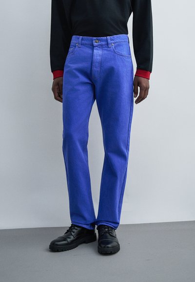 Persona che indossa jeans blu vivace a gamba dritta, scarpe nere e una maglia nera a maniche lunghe con polsini rossi su sfondo semplice.