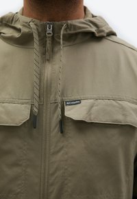 Κοντινή λήψη ενός χακί μπουφάν με φερμουάρ μπροστά, δύο τσέπες στο στήθος με καπάκι, κορδόνια και ετικέτα της μάρκας Columbia στην δεξιά τσέπη.