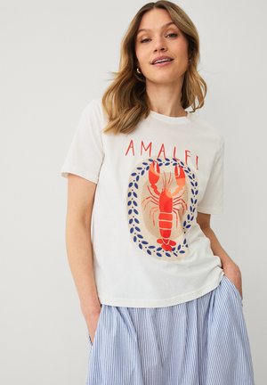 Kvinde iført en hvid T-shirt med rød hummert grafik og "Amalfi" tekst, kombineret med et blåt og hvidt stribet skørt.
