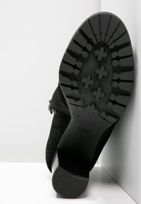 Bottines en suede noir avec une semelle en caoutchouc texturée, présentant un motif de bande de roulement et un logo en relief. La vue en angle met en valeur le design et le matériau.