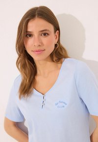 T-shirt en coton bleu clair, à manches courtes, avec un col en V et trois boutons, comportant un texte brodé "Los Palmeras CLUB" en bleu plus foncé.