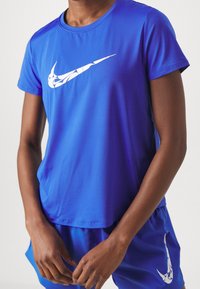 Persona vistiendo una camiseta atlética azul y pantalones cortos con logotipos abstractos en blanco de Nike en el pecho y el muslo.
