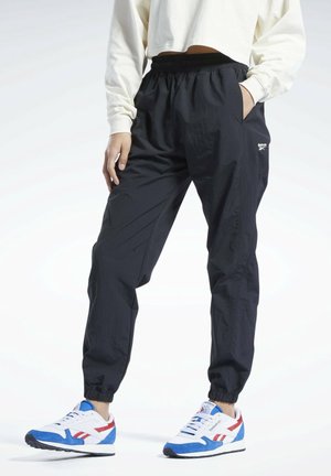 Personne portant un pantalon de jogging noir à taille élastique et des baskets Reebok blanches, bleues et rouges, debout avec une main dans la poche et une main sur le côté.