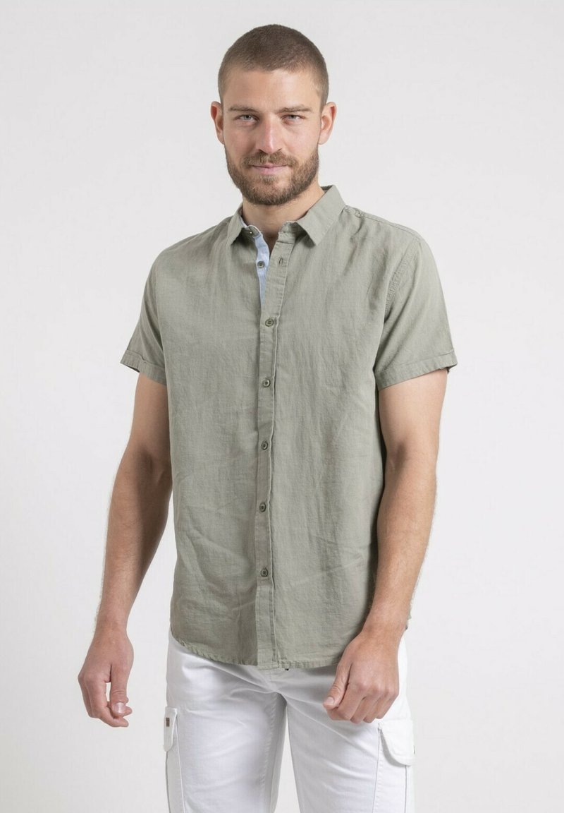 Chemisette à manches courtes de couleur vert olive en lin, avec un col, une fermeture à boutons et une texture subtile. Associée à un pantalon blanc.
