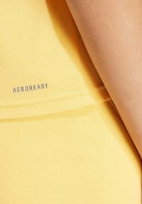 Top atlético amarillo hecho de tejido texturizado, con un logo "AEROREADY" visible en gris claro cerca del dobladillo.