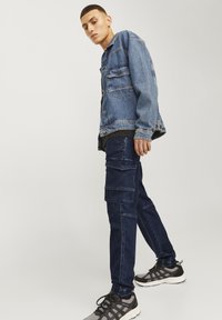 Chaqueta de mezclilla, azul, con dos bolsillos en el pecho y cierre de botón. Pantalones cargo oscuros de algodón, con bolsillos laterales. Zapatillas deportivas negras y grises.