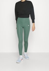 Sudadera negra recortada con ajuste relajado, combinada con leggings verdes de talle alto que presentan un detalle de logo. Zapatillas deportivas de color azul claro.