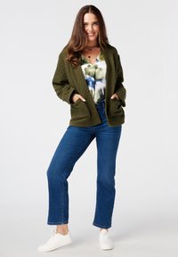 Gequilted olijfgroene jas met een relaxte pasvorm, voorzakken, gelaagd over een top met bloemenpatroon, gecombineerd met blauwe jeans en witte sneakers.