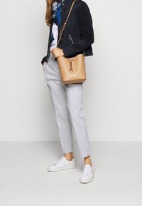 Béžová kožená kabela ve tvaru kyblíku s utahovací šňůrkou, námořnická semišová bunda, bílý tričko, šedo-pruhované kalhoty a bílé tenisky.