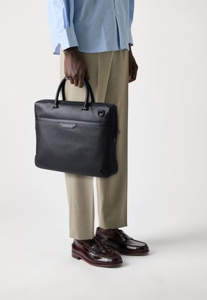 Attaché-case en cuir noir avec deux poignées supérieures, une fermeture éclair et un logo discret. Il a une texture lisse et une forme rectangulaire.