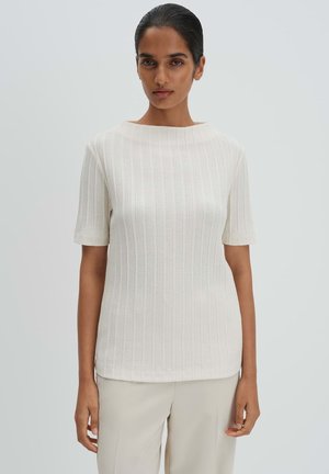 Frau trägt ein geripptes, kurzärmeliges, elfenbeinfarbenes Strickoberteil und hellbeige Hosen vor einem einfarbigen hellgrauen Hintergrund.