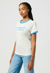 Camiseta de manga corta blanca con ribete azul, que presenta el texto "Una Leyenda en Jeans" en azul claro en la parte frontal. Material de algodón suave.