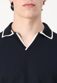 Mornarska polo majica s rebrastom teksturom, s V-izrezom i kontrastnim bijelim obrubom ovratnika. Akcenti uključuju vertikalno rebranje.