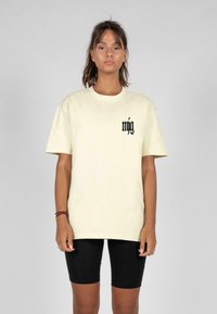Lys gul, løstsiddende T-shirt lavet af bomuld, med et sort logo på venstre bryst; parret med sorte cykelshorts.