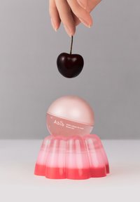 Masque pour les lèvres au collagène PDRN Abib dans un contenant rond et rose, posé sur une base translucide semblable à de la gelée. Une cerise est suspendue au-dessus du produit.