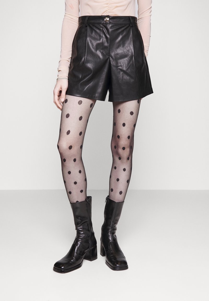 Personne portant un short en cuir noir, des collants transparents à pois et des bottes noires à talons mi-mollet, debout devant un fond uni.