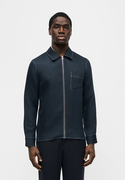 Άνδρας που φοράει σκούρα μπλε μπλούζα με φερμουάρ και λευκή ραφή, με τσέπη στο στήθος, στέκεται μπροστά σε απλό ανοιχτό φόντο.