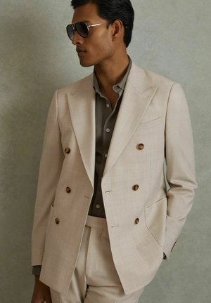 Veste de costume beige à double boutonnage avec des boutons marron, associée à une chemise boutonnée beige clair, et portant des lunettes de soleil noires.