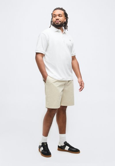 Άνδρας με γένια και ντερέκλες που φοράει λευκό πόλο, μπεζ σορτς, λευκές κάλτσες και μαύρα αθλητικά παπούτσια, στέκεται με τα χέρια στις τσέπες.