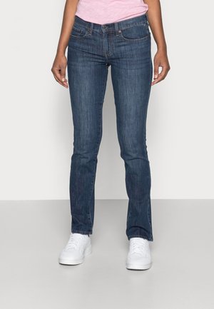 Kvinde iført tætsiddende mørkeblå jeans, hvide sneakers og en lyserød skjorte, stående mod en ensfarvet lys baggrund.