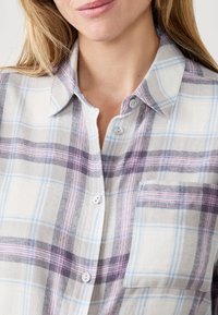 Koszula flanelowa w kratę w białym, szarym i lawendowym kolorze. Posiada kołnierzyk zapinany na guziki, kieszonkę z przodu oraz miękki, teksturowany materiał.