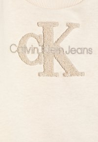 Camicia di cotone beige chiaro con logo ricamato "CK" in marrone chiaro testurizzato, e "Calvin Klein Jeans" ricamato sotto in un elegante grigio.