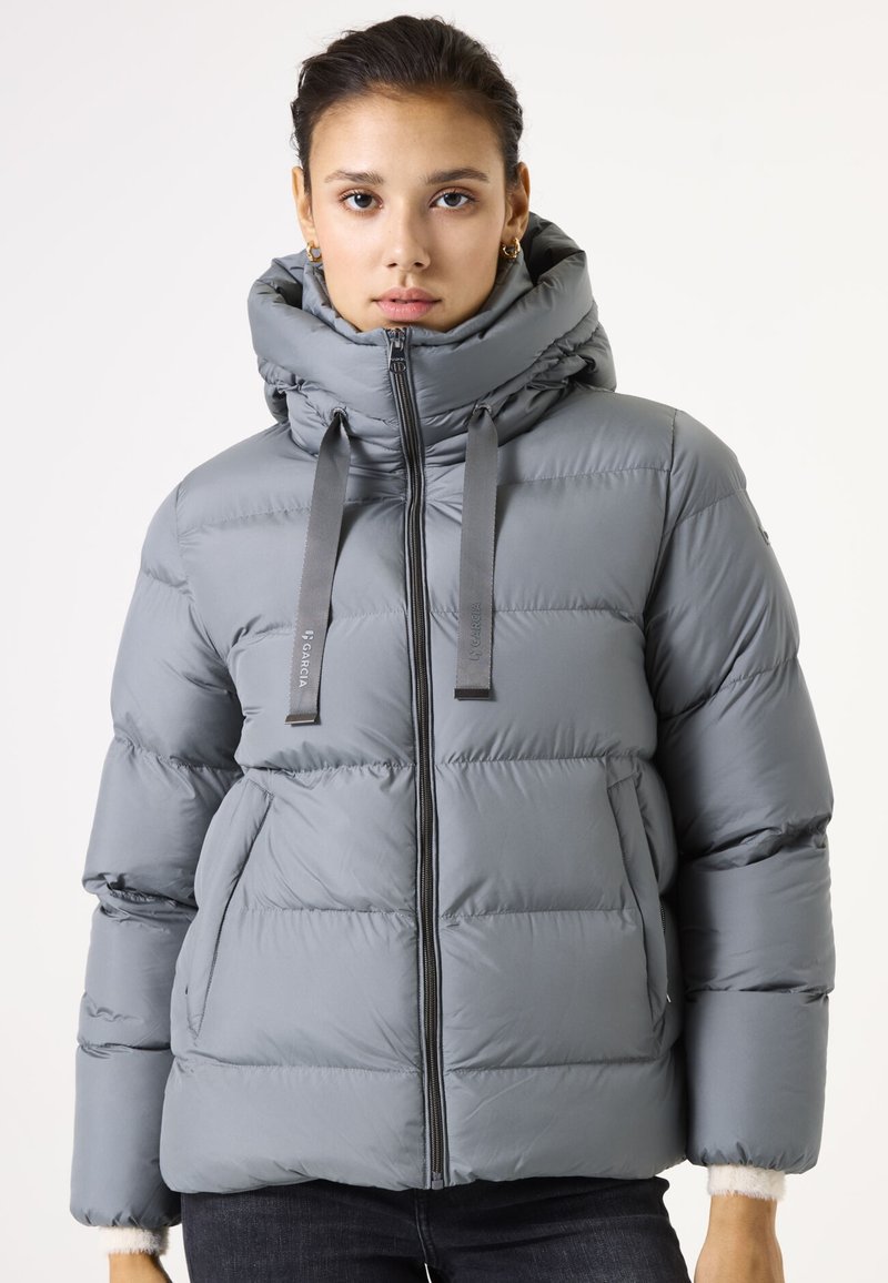 Grijze puffy jas met een hoge kraag, ritssluiting en lurex accenten. Heeft een boxy vorm en twee voorzakken. Zachte, gladde textuur.