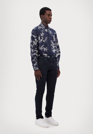 Chemise marine avec motif floral blanc, à manches longues, se fermant par des boutons, associée à un jean sombre et des baskets blanches. Texture de tissu lisse.