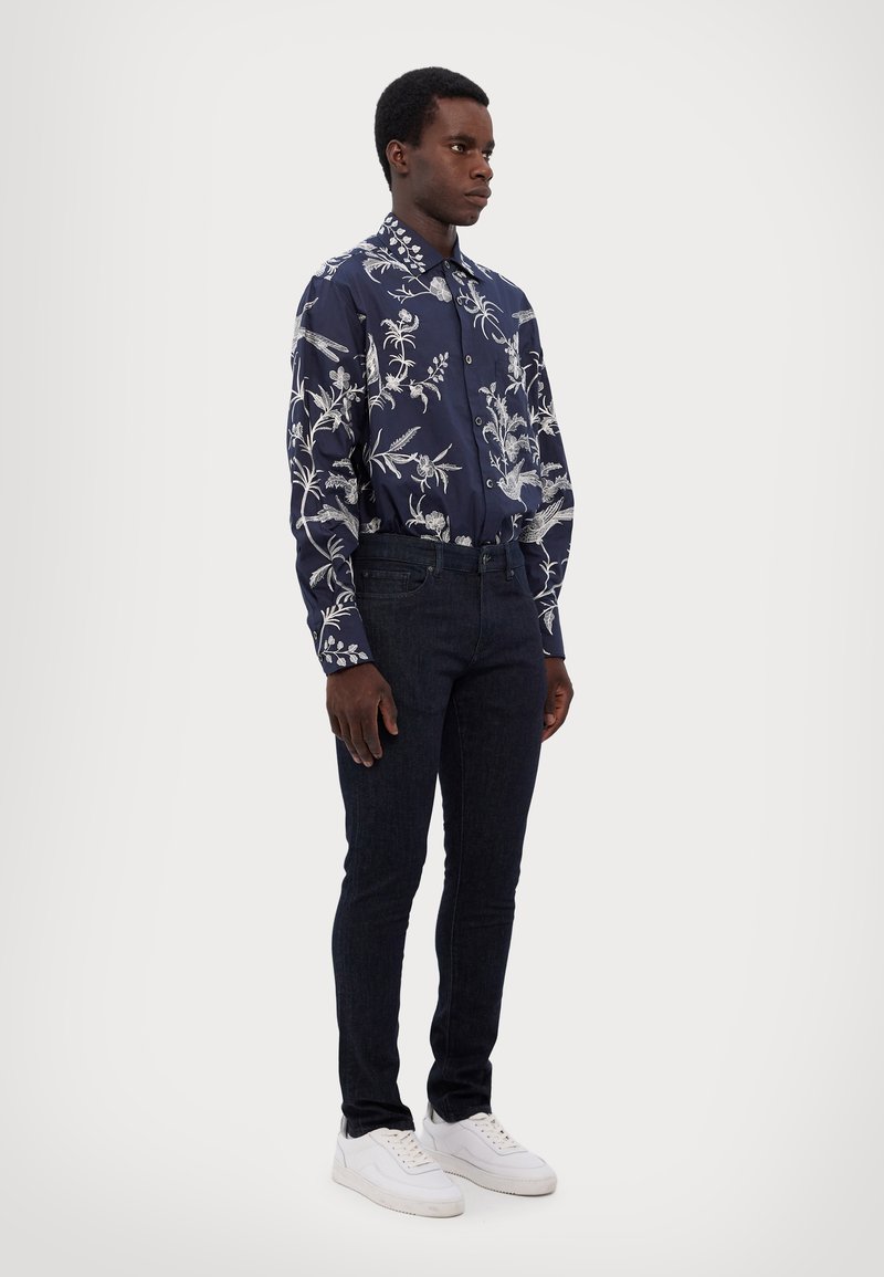 Camisa de marina con patrón floral blanco, de mangas largas, con botones en la parte delantera, combinada con jeans oscuros y zapatillas blancas. Textura de tela suave.