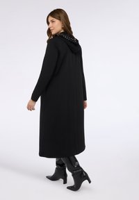 Mustapukuinen pitkä takki hupulla, jossa on niittiyksityiskohtia, istuvat hihat ja sileä pinta, yhdistettynä polvea ylempiin kiiltävän mustiin saappaisiin.