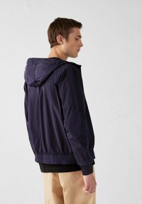 Giacca leggera blu navy con cappuccio, polsini e orlo elasticizzati, e superficie liscia. Progettata per una vestibilità rilassata, mostrata da dietro.