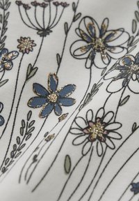 Vitt tyg med broderade blommönster i blått, rosa och guld, med ömtåliga stjälkar och blad som omger designen.