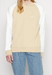Sudadera beige con mangas raglán blancas, cuello redondo y pequeño logo en el pecho. Fabricada en un material suave con puños y dobladillo acanalados.