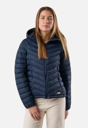 Junge Frau mit langen, glatten Haaren, die eine dunkelblaue gesteppte Kapuzenjacke mit Reißverschluss und hellbeige Hose trägt, vor einem einfarbigen Hintergrund stehend.