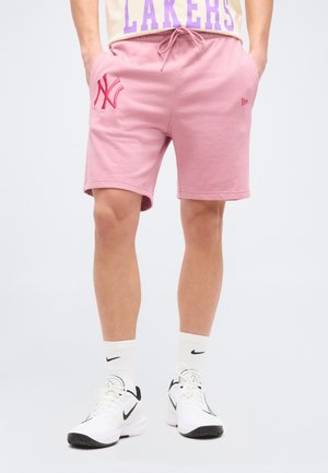 Άτομο που φοράει ροζ σορτς New York Yankees, λευκές κάλτσες Nike και λευκά αθλητικά παπούτσια Nike, στέκεται με τα χέρια στις τσέπες.