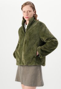 Vrouw in een groene nepbontjas met de handen in de zakken en een geruite rok, staand tegen een witte achtergrond.