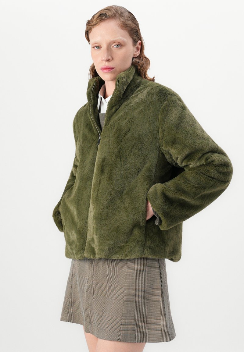 Frau trägt eine grüne Kunstfelljacke mit Händen in den Taschen und einem karierten Rock, stehend vor einem weißen Hintergrund.
