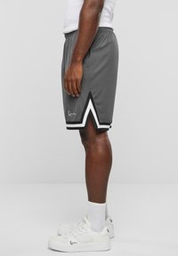 Homme portant un short de basket gris avec un liseré chevron blanc et noir et des baskets blanches avec un logo assorti, debout de côté sur un fond uni.