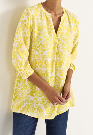 Woman wearing a yellow and white floral patterned tunic with 3/4 sleeves and dark blue jeans, hands clasped in front.