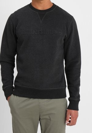 Mężczyzna ubrany w czarną, długą bluzę z okrągłym dekoltem i jasne spodnie oliwkowe, stoi z jedną ręką w kieszeni na białym tle.