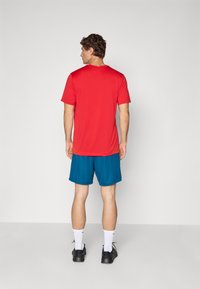 Red short-sleeve athletic shirt, paired with blue shorts, wearing white socks and black shoes. Model shown from the back, with curly hair.