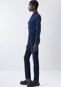 Mono de mezclilla azul oscuro con dos bolsillos en el pecho, diseño ajustado, mangas largas y cierres de botón, combinado con zapatos de plataforma negros.